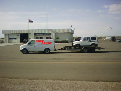 1987 Suzuki Samurai - A white Suzuki Samurai is transported on a trailer pulled by a white van that has 'FREE CANDY' written on its side.