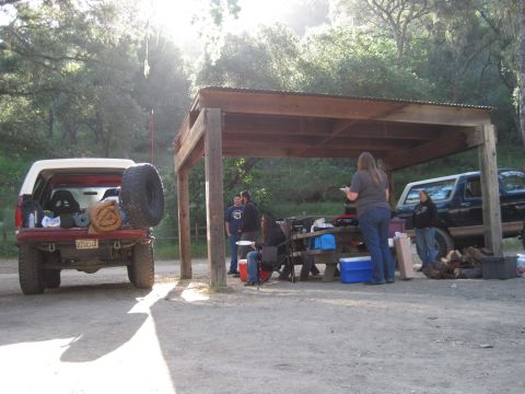 1989 Ford Bronco - People are gathered at a picnic shelter next to a red SUV and a black SUV in a wooded area.