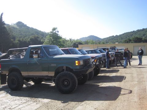 1989 Ford Bronco - A line of modified trucks and SUVs are parked outdoors on a sunny day.