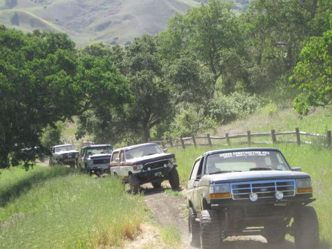 1989 Ford Bronco - A convoy of Ford trucks and Broncos navigates a dusty, rural path lined with trees and a wooden fence.