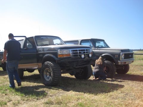 1989 Ford Bronco - Two modified SUVs, a Ford Bronco and a Chevrolet K5 Blazer, are parked on a grassy field under a clear blue sky.