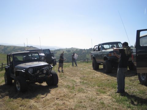 1989 Ford Bronco - A group of off-road vehicles, including a lifted green 1989 Ford Bronco, are parked on a grassy hillside under a clear blue sky.