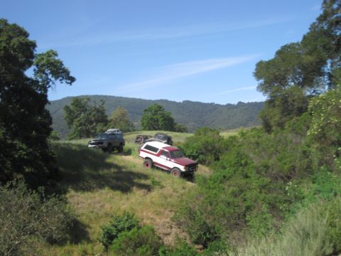 1989 Ford Bronco - Multiple off-road vehicles are parked on a grassy hillside under a clear blue sky.