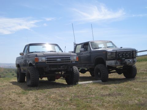 1989 Ford Bronco - Two lifted Ford trucks, a black one on the left and a dark gray one on the right, are parked side-by-side on a grassy hill.