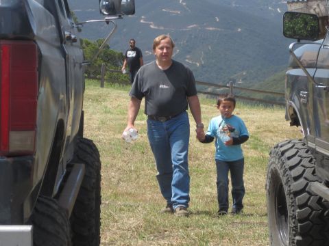 1989 Ford Bronco - A man and boy walk between two large trucks on a grassy hillside with a distant view of winding roads.