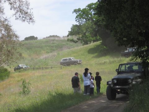 1989 Ford Bronco - Several off-road vehicles are parked and driving on a grassy hill with a group of people observing.