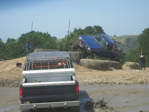 1989 Ford Bronco - A blue pickup truck is precariously balanced on large tires, while another truck navigates muddy terrain.