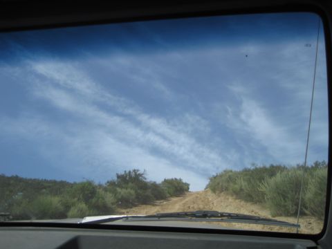 1989 Ford Bronco - View from inside a vehicle looking up a dirt road with scrub brush under a cloudy blue sky.