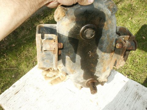 1966 Ford Bronco - A greasy, rust-streaked cast-iron transfer case or gearbox housing stood upright on a white-painted plank in the grass, steadied by a bare…