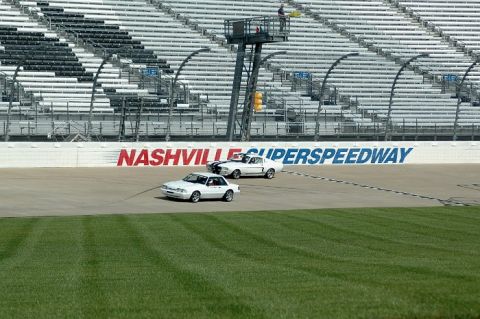 2007 Ford Mustang - Two white Ford Mustangs race on a track with grandstands in the background and 'NASHVILLE SUPERSSPEEDWAY' signage.