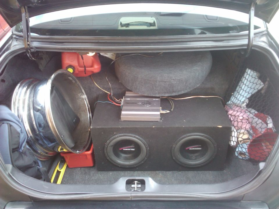 Car trunk interior with a spare tire, gas can, large subwoofer box, amplifier, and a chrome rim