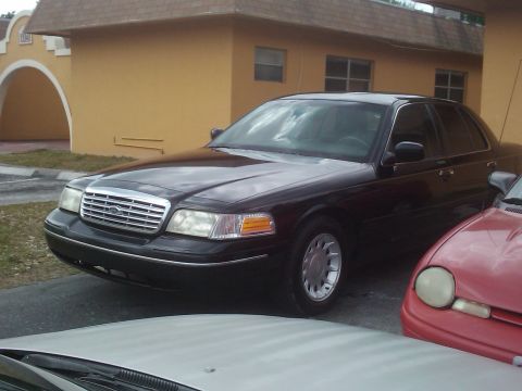 2001 Ford Crown Victoria - A dark-colored sedan is parked in front of a yellow building, with parts of two other vehicles visible.