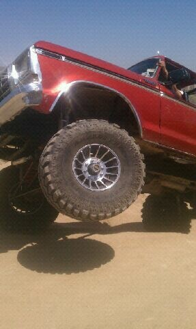 1978 Ford Bronco - A red 1978 Ford Bronco is captured from a low angle, its lifted suspension and large off-road tires against a sandy backdrop.