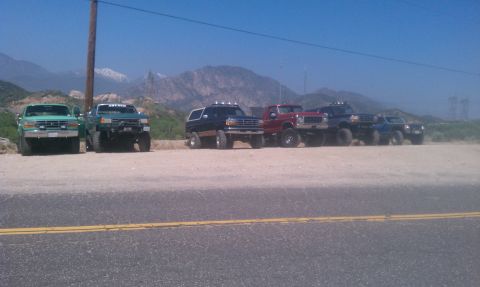 1978 Ford Bronco - A line of lifted Ford Broncos and trucks are parked on a dirt lot with mountains in the background.
