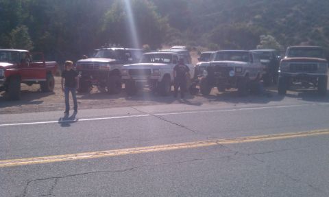 1978 Ford Bronco - A collection of off-road vehicles are lined up on a roadside, with a mountainous backdrop under a bright sun.