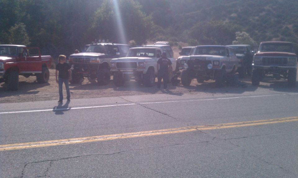 Several lifted trucks and SUVs parked on the side of a road with hills in the background. Two people are standing near the vehicles