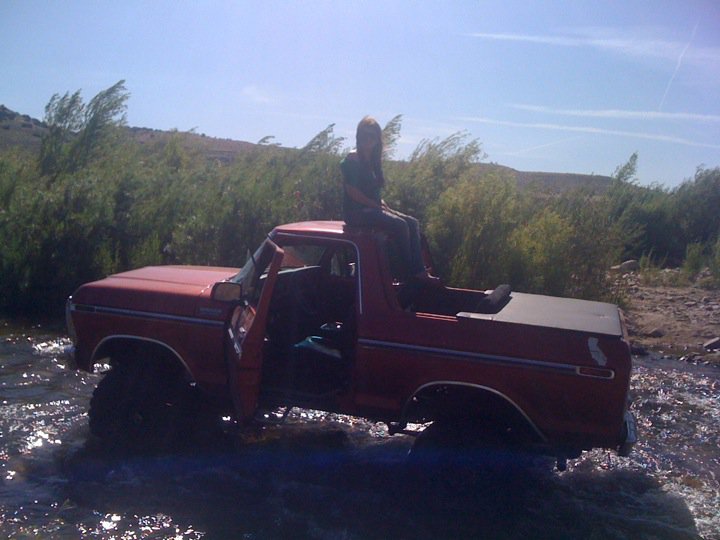 1978 Ford Bronco parked in a stream with the driver's door open and a person sitting on the roof