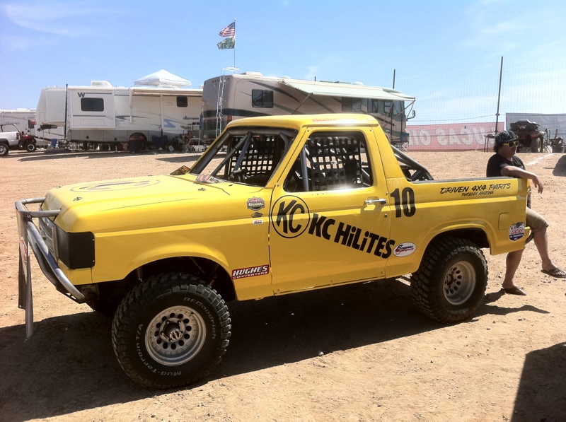 Yellow off-road truck with KC HiLiTes decals and a roll cage, parked in a sandy lot with RVs behind it