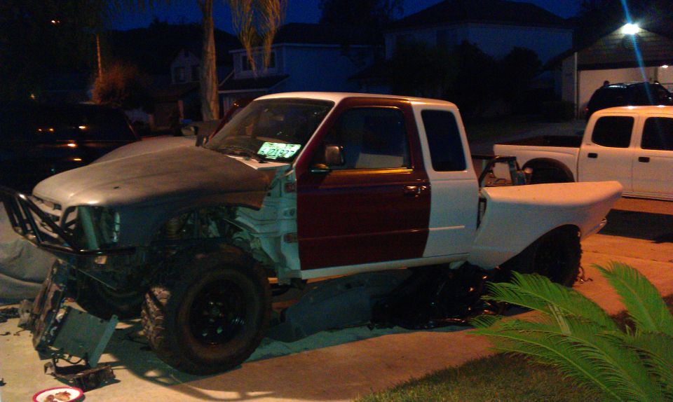 Truck with front end disassembled, primer hood, maroon driver door, and white passenger side and bed. Another white truck is parked behind it