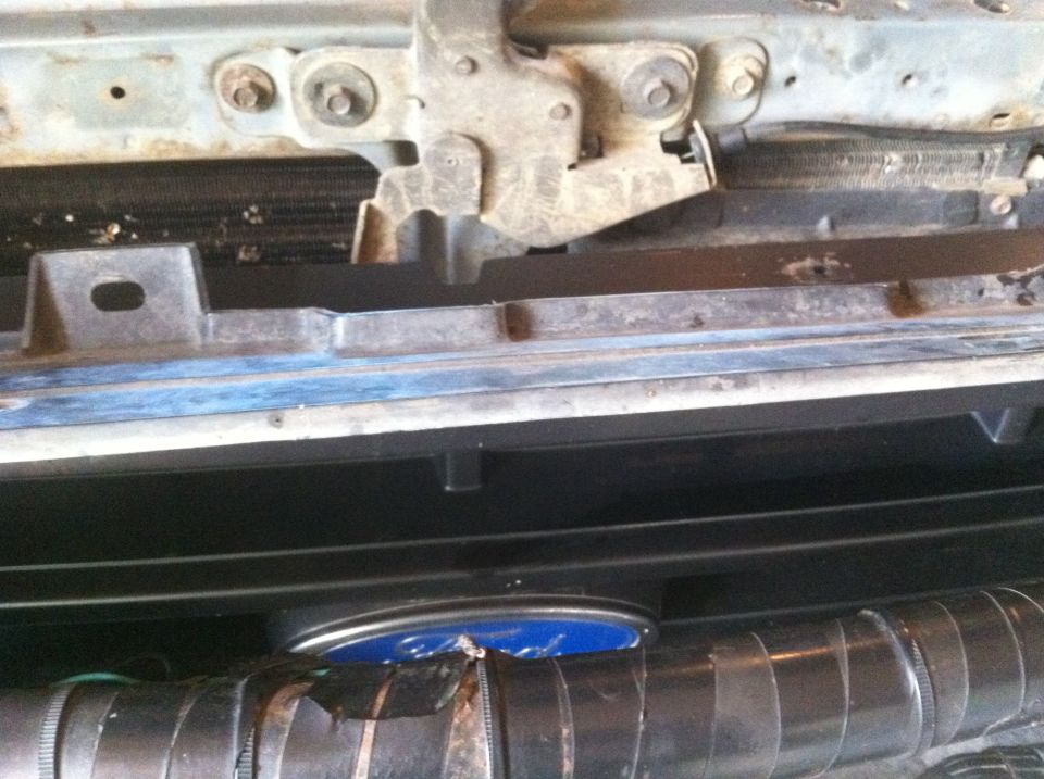 Close-up of a vehicle's front end, showing the hood latch mechanism and part of the grille. A blue emblem is visible