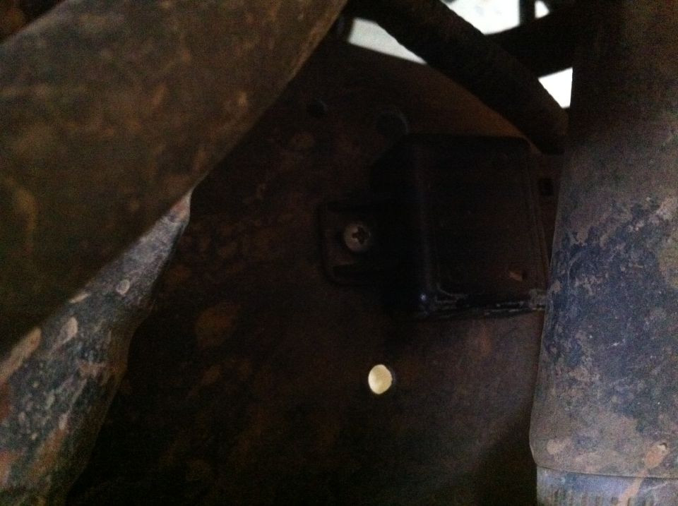 Underside of a dark vehicle part with a screw and a small hole, surrounded by metal pipes