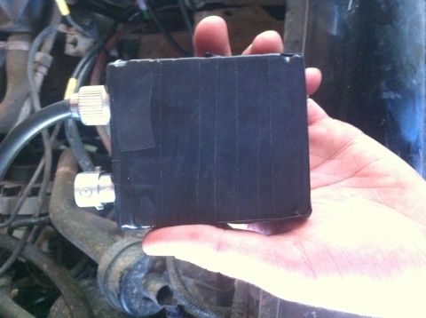 1990 Ford Bronco - A person holds a black, tape-wrapped electronic component with metal connectors in front of a vehicle's engine compartment.