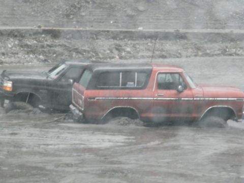 1978 Ford Bronco - Two vehicles, a red 1978 Ford Bronco and a black truck, are stuck in muddy terrain.