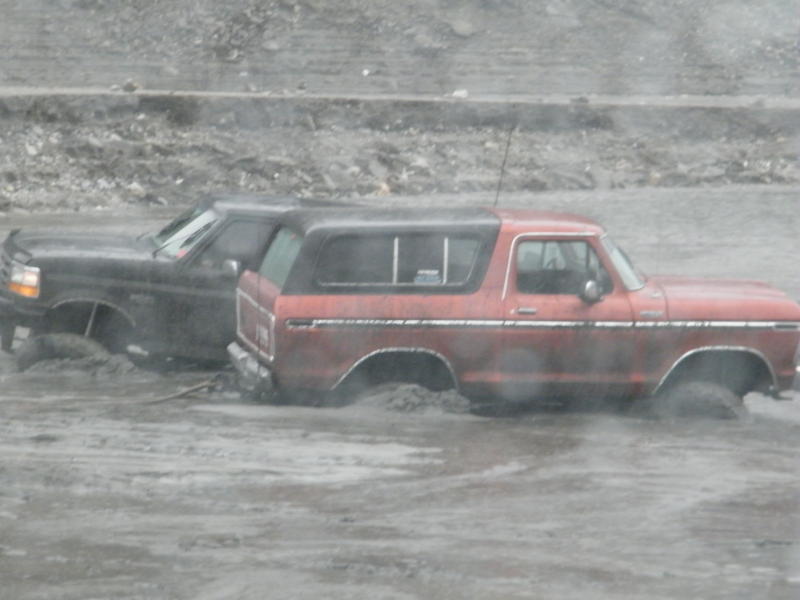 Red 1978 Ford Bronco side view stuck in mud next to a black truck