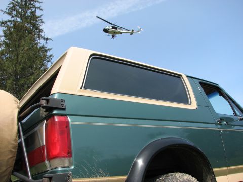 1990 Ford Bronco - A green Ford Bronco with a tan roof trim is viewed from the rear quarter panel, with a helicopter flying in the sky.