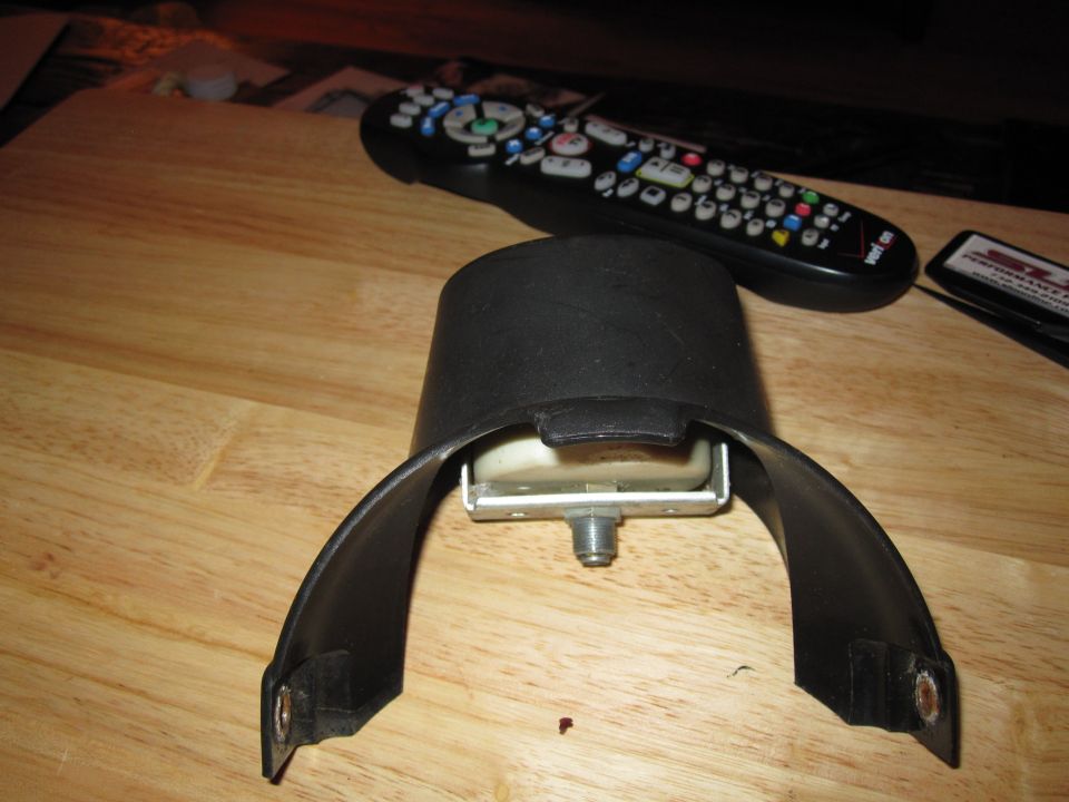 Seatbelt buckle mechanism with black plastic housing and metal components on a wooden surface. Remote control and a small card are visible in the background