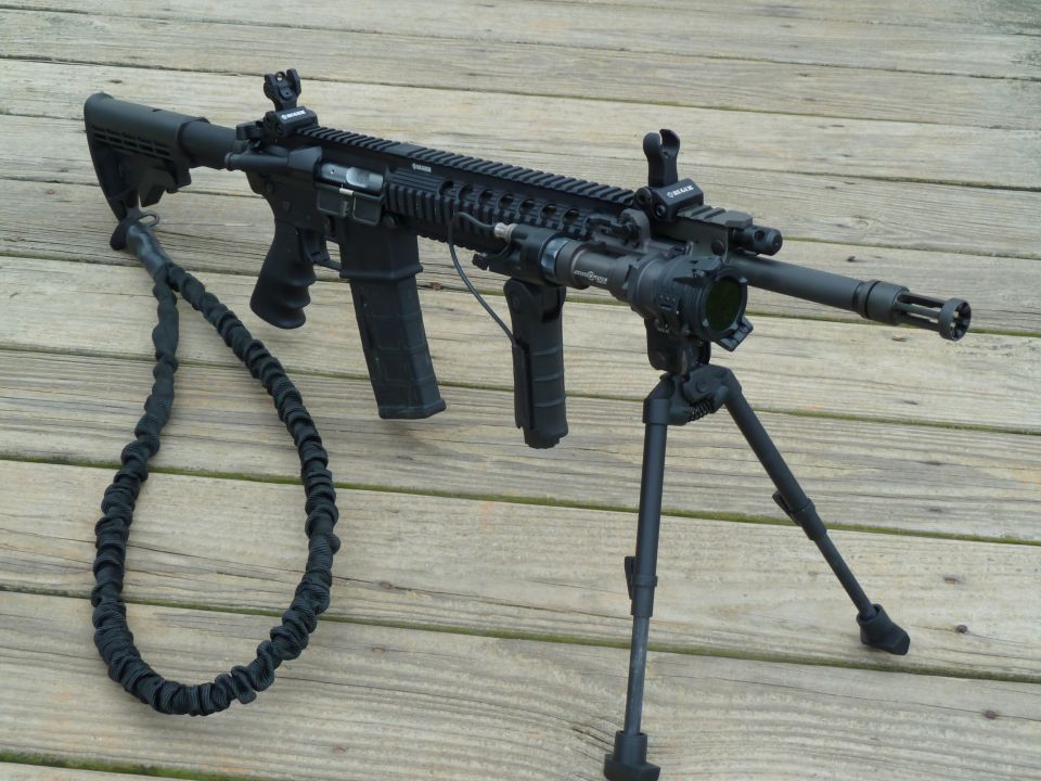 Black assault rifle with bipod, tactical light, and sling on a wooden deck