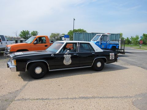 1991 Ford LTD Crown Victoria - black-and-white Ford police sedan numbered 45 with a star-style door shield parked on gravel near an orange pickup.