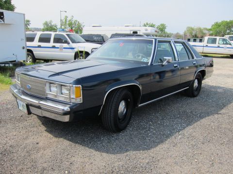 1991 Ford LTD Crown Victoria - dark blue Ford four-door sedan with a window tag parked on a gravel lot among other vehicles under a hazy sky.