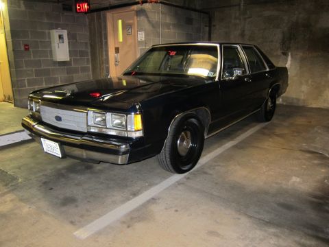 1991 Ford LTD Crown Victoria - black Ford four-door sedan with a red dashboard light parked in a concrete parking garage near an exit door.