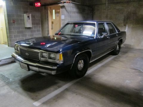 1991 Ford LTD Crown Victoria - dark blue Ford four-door sedan with a window tag parked in a dim concrete parking garage near an exit sign.