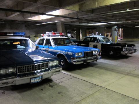1991 Ford LTD Crown Victoria - blue Dodge sedan marked 'POLICE' with roof lights parked among other police cruisers inside a concrete garage.