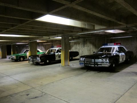 1991 Ford LTD Crown Victoria - Three classic police cars, including a black-and-white highway patrol sedan and a green sheriff car, parked in a dim concrete parking struc…