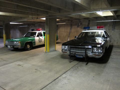 1991 Ford LTD Crown Victoria - green-and-white sheriff sedan beside a black-and-white police car with a push bar, parked in a dim concrete garage.