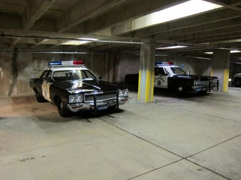 1991 Ford LTD Crown Victoria - Two black-and-white vintage police sedans with roof light bars parked diagonally in a worn concrete parking structure.