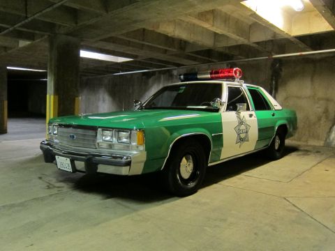 1991 Ford LTD Crown Victoria - green-and-white Ford police sedan with a red-and-blue roof light bar parked in a dim concrete parking structure.
