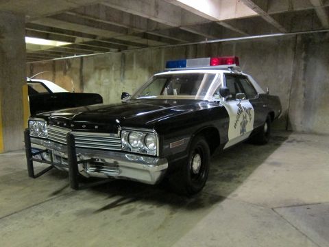 1991 Ford LTD Crown Victoria - black-and-white Dodge Highway Patrol sedan with push bar parked in a concrete garage next to a white sedan with an open hood.