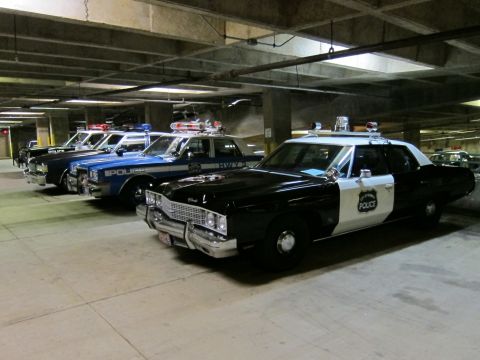 1991 Ford LTD Crown Victoria - row of vintage police cars, including black-and-white Police sedans and a blue cruiser, parked in a dim concrete parking structure.