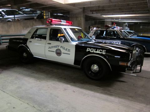 1991 Ford LTD Crown Victoria - black-and-white sedan marked Jaffrey Police with a red roof light bar parked in a dim concrete garage beside another police car.