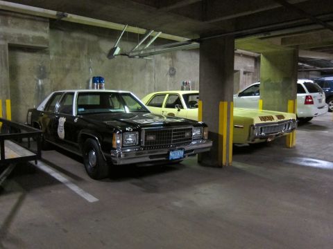 1991 Ford LTD Crown Victoria - black-and-white police sedan parked beside a yellow State Police sedan in a dim concrete parking structure.