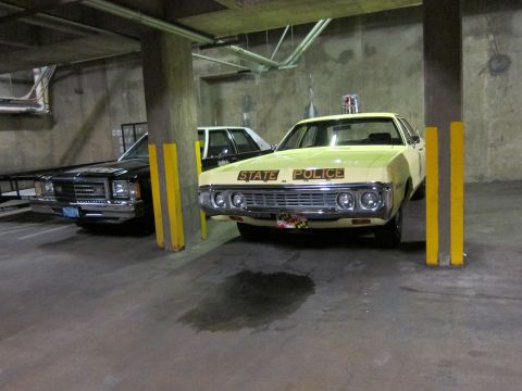 1991 Ford LTD Crown Victoria - yellow sedan marked State Police with a roof beacon parked next to a black car in a dim concrete parking structure.