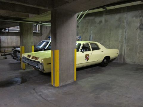 1991 Ford LTD Crown Victoria - yellow State Police sedan and a black police car parked on either side of a concrete support pillar in a dim garage.