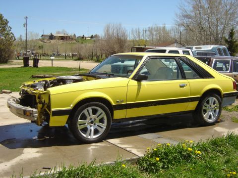 1979 Ford Mustang - A yellow coupe sits in a driveway with its hood up and front bumper missing, indicating it is undergoing repairs.