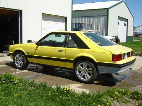 1979 Ford Mustang - A yellow 1979 Ford Mustang coupe sits in front of a garage, showing its side profile.