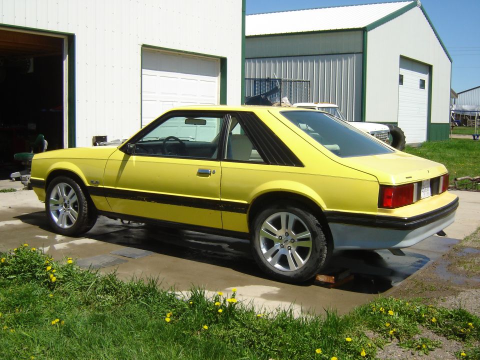 Yellow 1979 Ford Mustang coupe parked outside a garage, viewed from the side