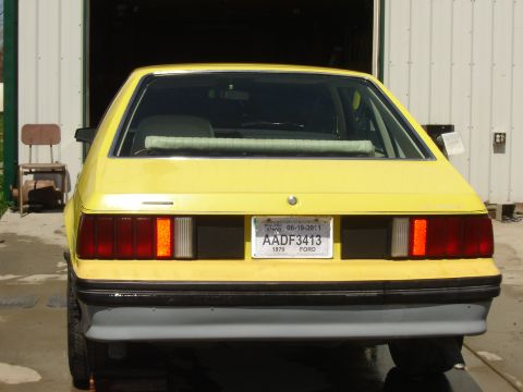1979 Ford Mustang - Yellow 1979 Ford Mustang parked in front of a garage, showing its rear end and license plate.